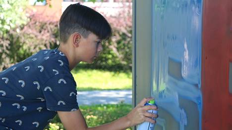 12-vuotias Luka Lausniemi maalaa tekstigraffitia Kaalisaaren padel-kenttien vieressä olevan muuntajakopin seinään. Lausniemi on aiemmin kokeillut spraymaaleilla maalaamista vain virtuaalitodellisuudessa.